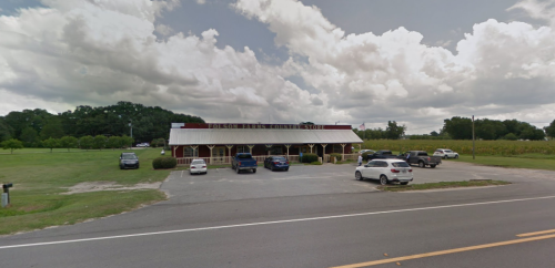 A country store with a wooden facade, surrounded by green fields and parked cars under a cloudy sky.