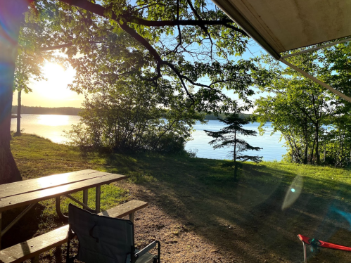 A serene lakeside view at sunset, with trees, a picnic table, and a camping chair in the foreground.
