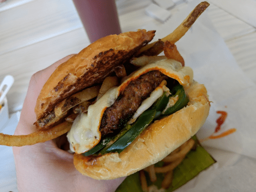 A hand holding a burger with a beef patty, melted cheese, jalapeños, and fries, with a drink in the background.