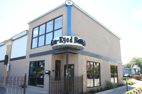 Exterior of a modern building with large windows and a sign reading "The Eyed Betty" in a vibrant neighborhood.