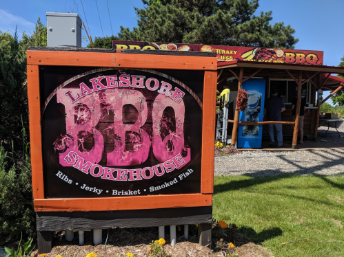 Sign for Lakeshore BBQ Smokehouse featuring ribs, jerky, brisket, and smoked fish, with a food stand in the background.