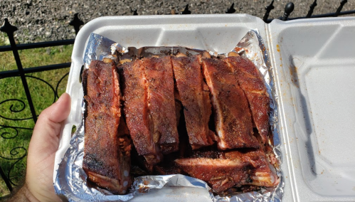 A hand holding a takeout container filled with grilled, seasoned ribs on a bed of foil.