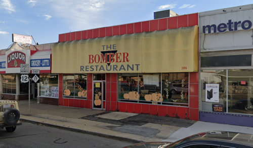 Exterior of The Bomber Restaurant, featuring a yellow awning and large windows, surrounded by other storefronts.