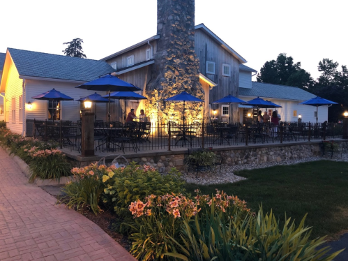 A cozy restaurant with outdoor seating under blue umbrellas, illuminated by warm lights at dusk. Lush flowers in the foreground.