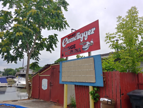Sign for "The Clamdigger Lounge & Pizzeria" with a red background, surrounded by greenery and boats in the background.