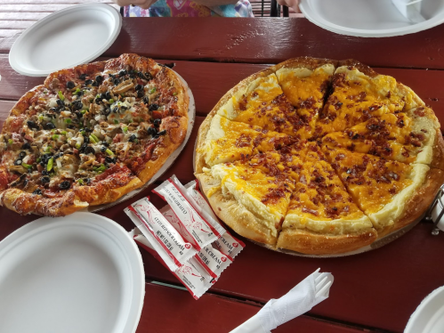 Two pizzas on a wooden table: one with vegetables and the other with cheese and bacon, accompanied by condiment packets.