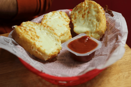 A basket with three cheesy bread pieces and a small container of marinara sauce on a wooden table.