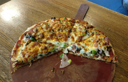 A partially sliced pizza on a wooden board, topped with cheese, green peppers, onions, and olives.