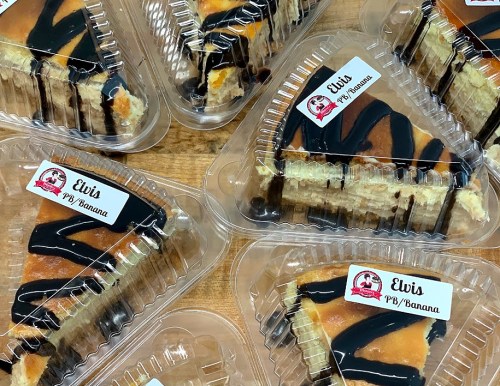 Slices of Elvis PB/Banana cheesecake in clear containers, drizzled with chocolate, arranged on a wooden surface.