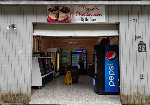 A cozy shop entrance with a sign for "Momo's Cheesecake By the Slice," featuring a Pepsi vending machine and a caution sign.