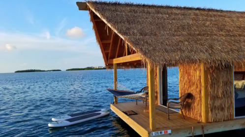 A thatched-roof hut on a dock over calm water, with a paddleboard and chairs nearby, surrounded by a scenic landscape.