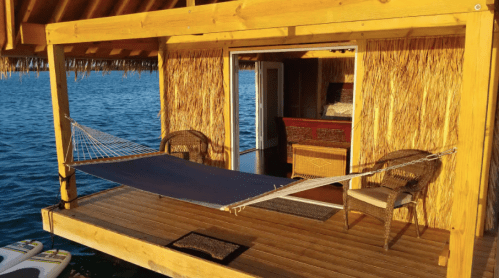 A cozy wooden deck with a hammock and chairs overlooking calm water, featuring a thatched-roof bungalow in the background.