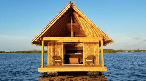 A wooden hut with a thatched roof on stilts over calm water, surrounded by greenery under a clear blue sky.