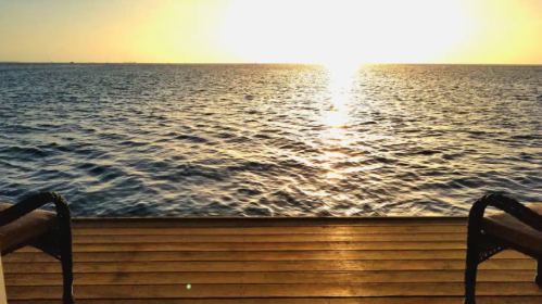 Sunset over a calm ocean, viewed from a wooden deck, with golden light reflecting on the water's surface.