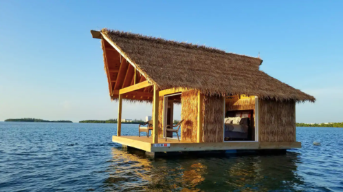 A thatched-roof hut on stilts in calm water, surrounded by blue skies and distant islands.