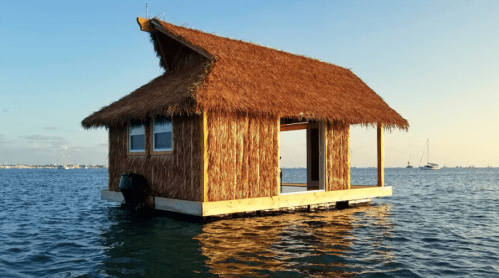 A thatched-roof floating hut on calm water, with an open doorway and a scenic backdrop of boats and sky.