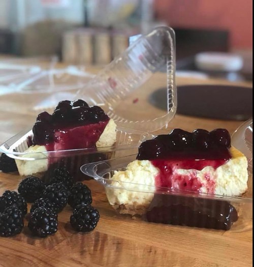 Two slices of cheesecake topped with berry sauce in clear plastic containers, surrounded by fresh blackberries.