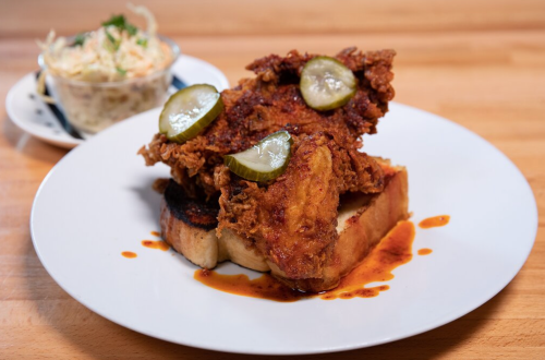 A plate featuring fried chicken on toast, topped with pickles, and a side of coleslaw in a small bowl.