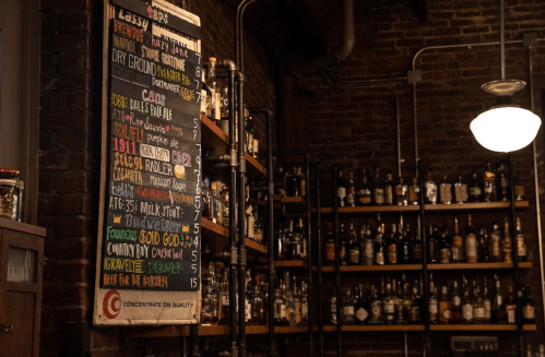 A chalkboard menu with drink options and prices, set against a backdrop of shelves filled with various liquor bottles.