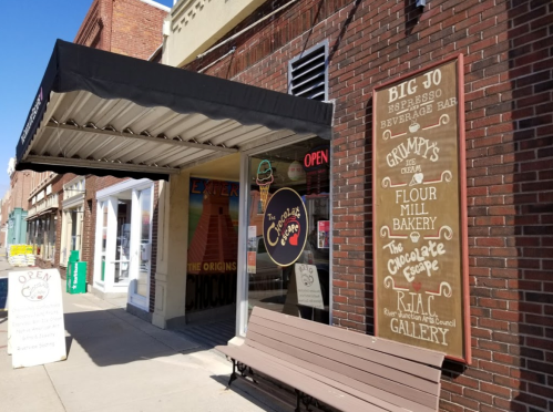 A storefront with a sign reading "The Chocolate Escape" and various nearby businesses, featuring an open sign and outdoor seating.