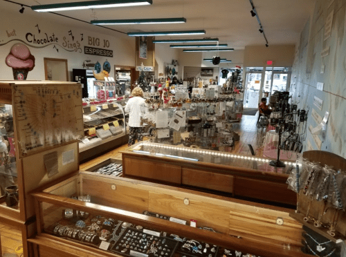 A cozy shop interior filled with jewelry displays, gifts, and a café area, with a customer browsing in the background.