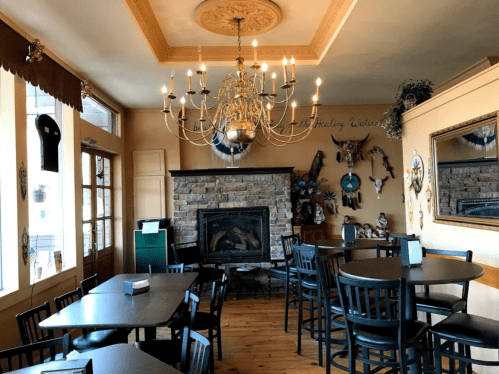 Cozy restaurant interior with wooden tables, black chairs, a stone fireplace, and a chandelier. Decor includes wall art.