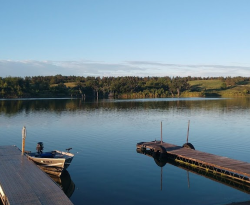 A calm lake scene with a small boat and a wooden dock, surrounded by lush greenery under a clear blue sky.