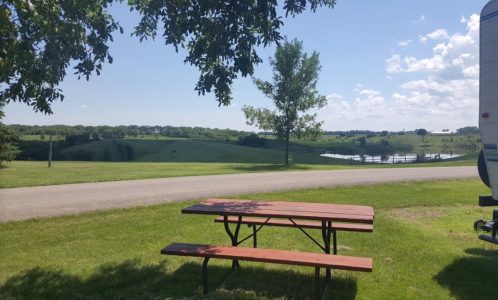 A picnic table sits on green grass by a road, overlooking a scenic landscape with hills and a pond under a blue sky.