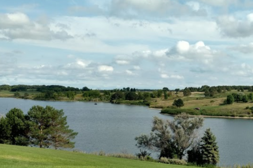 A serene landscape featuring a calm lake surrounded by green hills and scattered trees under a partly cloudy sky.
