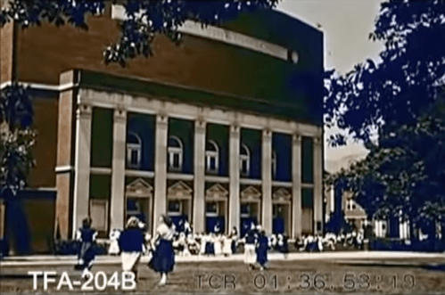 A historic building with columns, surrounded by people in formal attire, set in a park-like area.