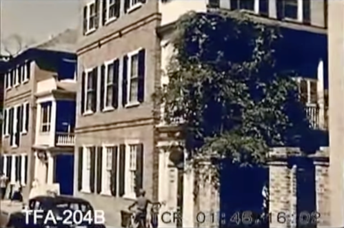 Historic buildings with ivy, a parked car, and people walking on a street in a vintage setting.