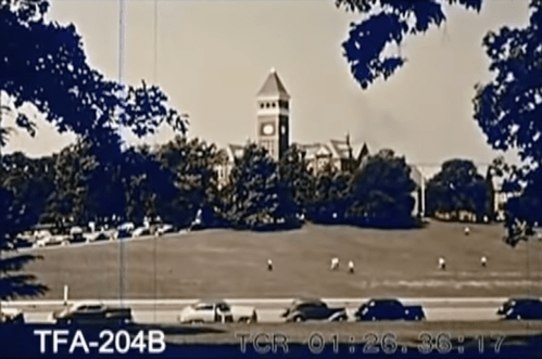 Historic black-and-white image of a clock tower on a campus, surrounded by trees and people on a grassy field.