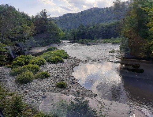 A serene river flows through a lush green landscape, surrounded by trees and mountains under a cloudy sky.