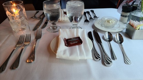 A dining table set with silverware, two glasses of water, a candle, and a plate with a chocolate on a napkin.