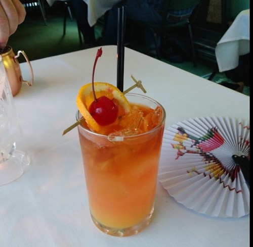 A colorful cocktail with ice, garnished with an orange slice and a cherry, served with a straw on a table.
