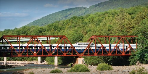 A train crosses a red metal bridge over a river, surrounded by lush green mountains and trees.