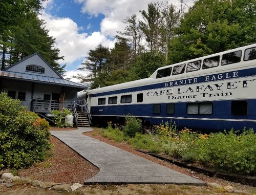 A vintage train car named "Café Lafayette" is parked next to a building, surrounded by greenery and flowers.