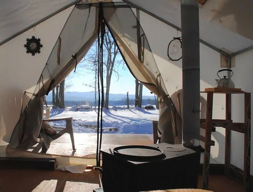 Interior of a tent with a view of snowy landscape and trees outside, featuring a stove and wooden furniture.