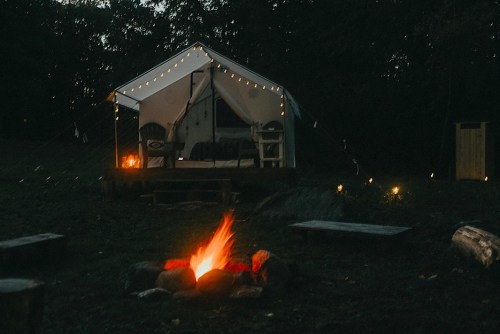 A cozy tent with string lights, surrounded by a campfire and rustic seating in a serene outdoor setting.