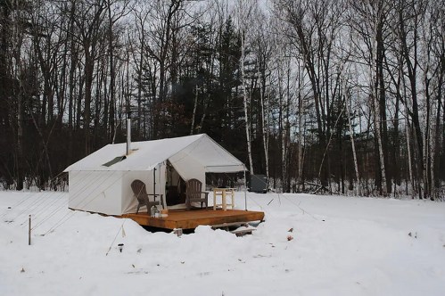 A cozy tent cabin on a snowy landscape, surrounded by trees, with wooden chairs on a deck.