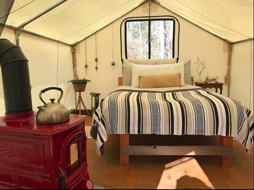Cozy glamping tent interior with a bed, decorative blanket, and a red stove, surrounded by natural light.