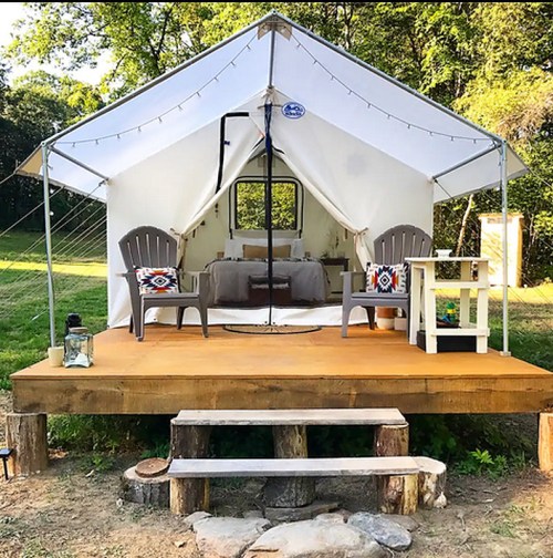 A cozy glamping tent on a wooden deck, featuring two chairs, a small table, and a lantern in a natural setting.