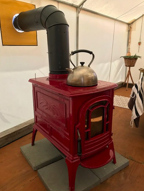 A red wood stove with a black chimney pipe and a kettle on top, set inside a tent with wooden flooring.