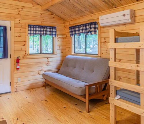 Cozy wooden cabin interior with a futon, bunk beds, and large windows, featuring a rustic design and natural light.