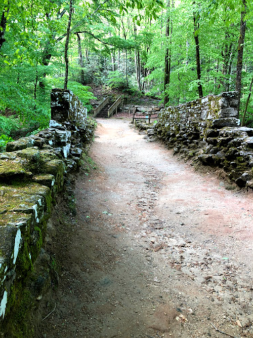 A stone bridge leads into a lush green forest, with a dirt path winding through trees and moss-covered rocks.