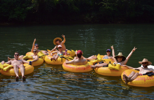 A group of people in yellow inner tubes floating on a river, enjoying a sunny day and posing with peace signs.
