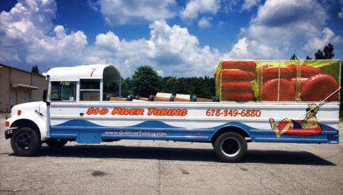 A bus converted for river tubing, featuring bright graphics and inflatable tubes on top, parked in a lot.