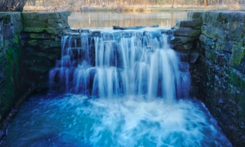 A serene waterfall cascades over stone steps into a calm, reflective pool surrounded by nature.