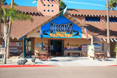 Exterior of Dillon's BBQ restaurant with a blue sign, picnic tables, and decorative pigs in front. Palm trees in the background.