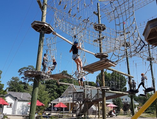A group of children navigating a rope course with nets and platforms in a sunny outdoor setting.